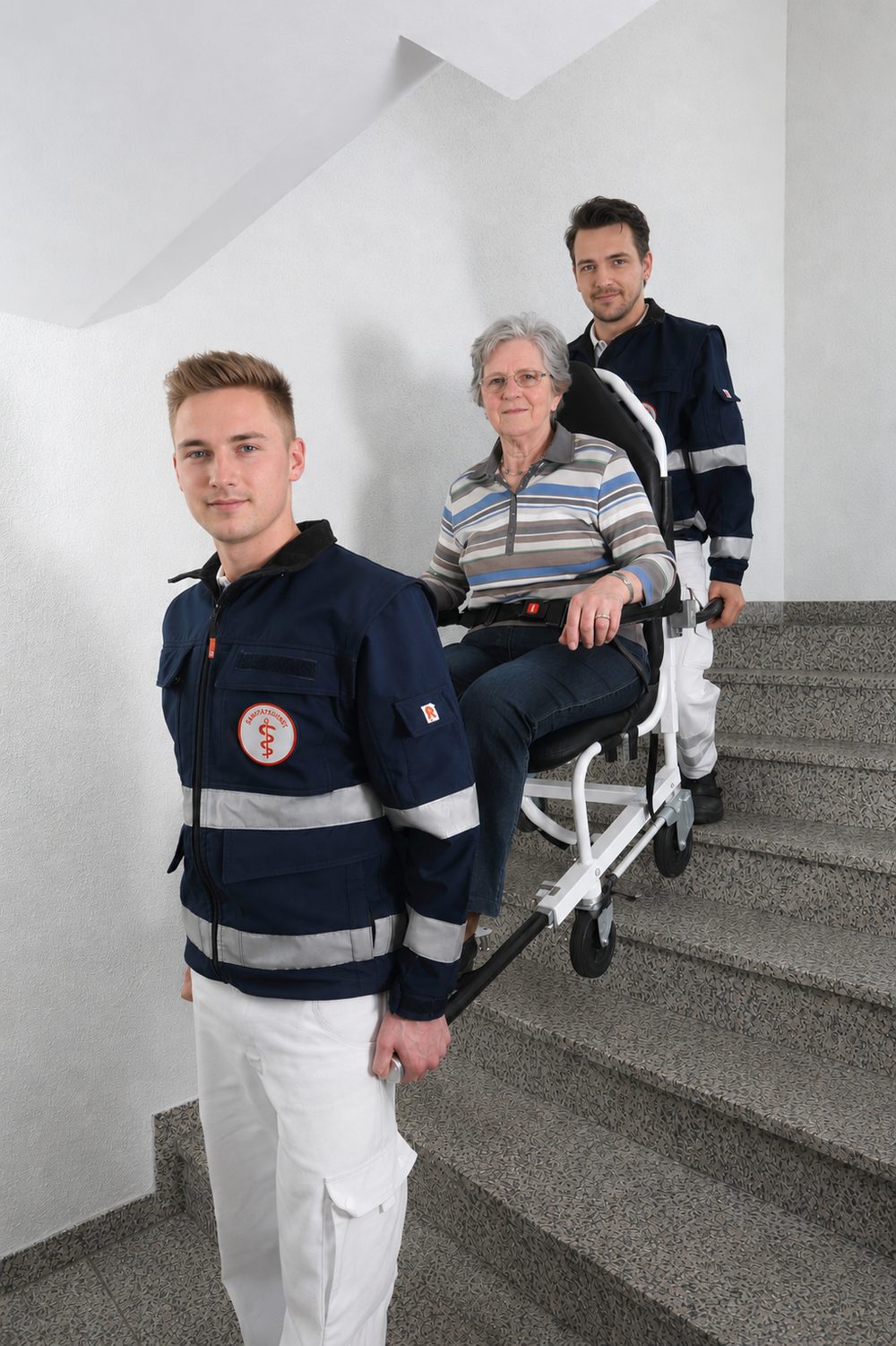 Zwei Medimobil-Mitarbeiter tragen eine Patientin im Tragestuhl sicher über eine Treppe im Hausflur